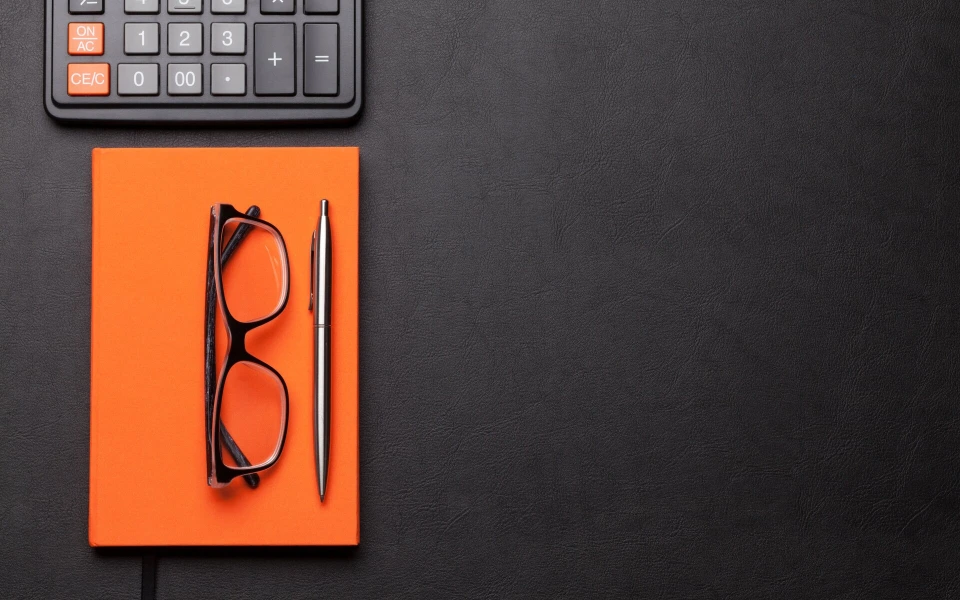 Taschenrechner, orangefarbenes Notizbuch und Brille auf schwarzem Schreibtisch
